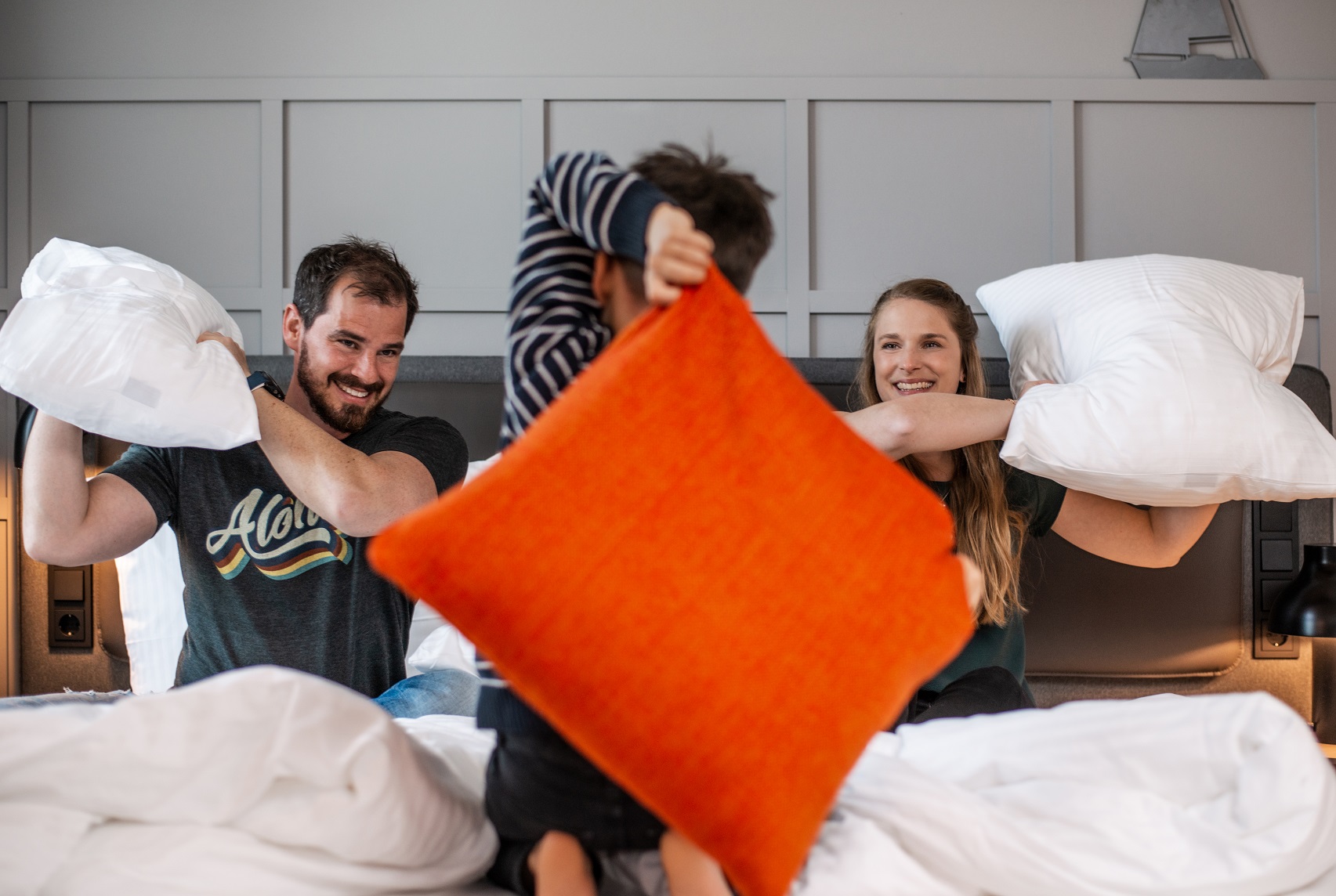 Familie hat Kissenschlacht auf Hotelbett, lachend, mit weißem und orangem Kissen, freundliche Atmosphäre.