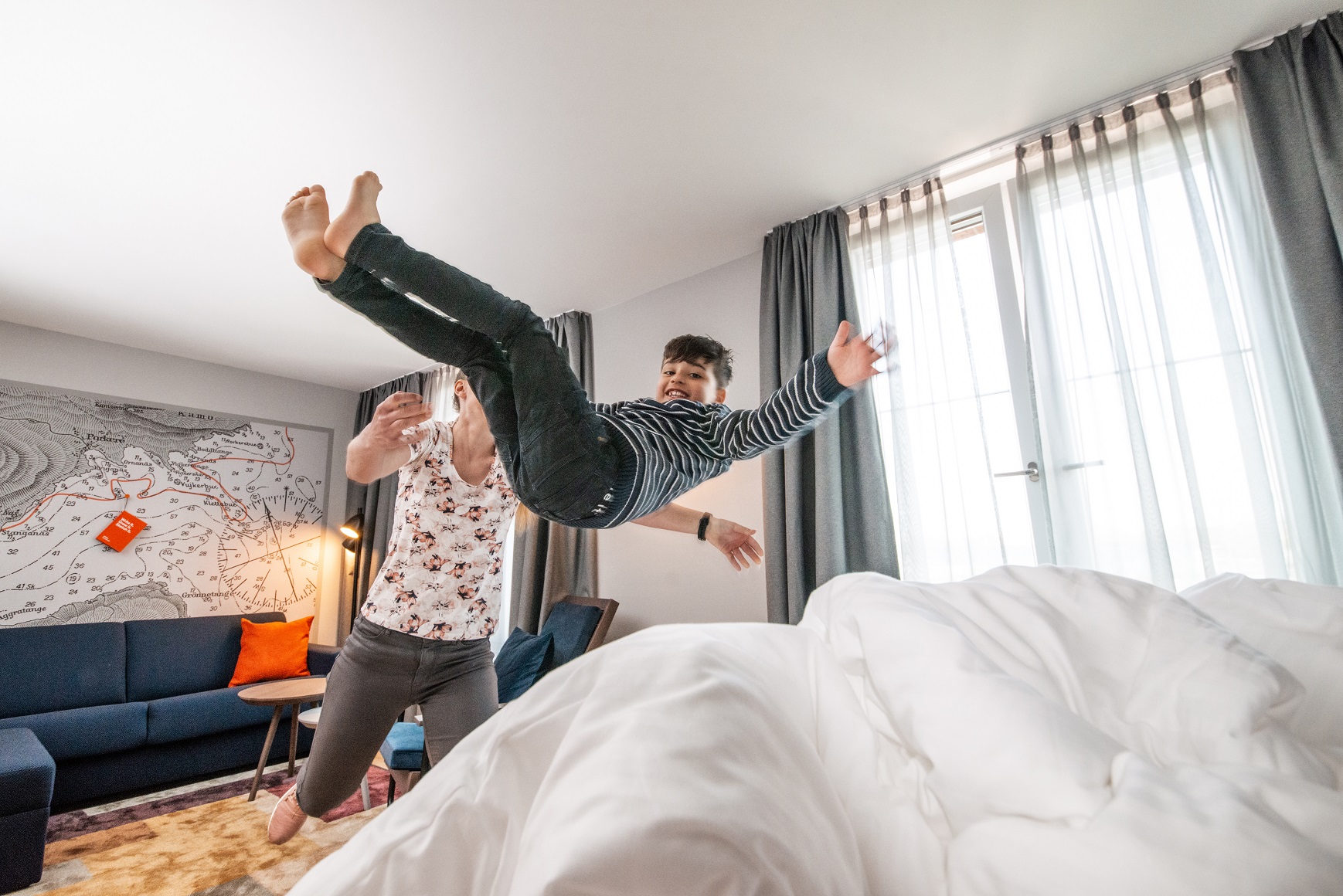 Ein Kind springt fröhlich auf einem Hotelbett, während eine erwachsene Person es auffängt.